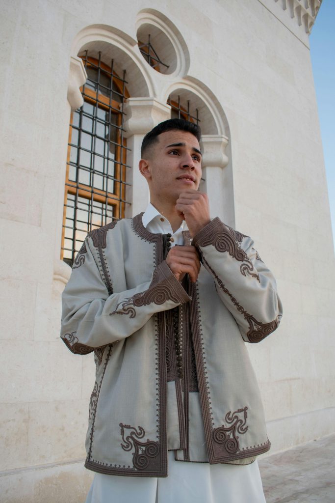 Portrait of a young man in traditional embroidered coat standing outdoors in front of an architectural structure.