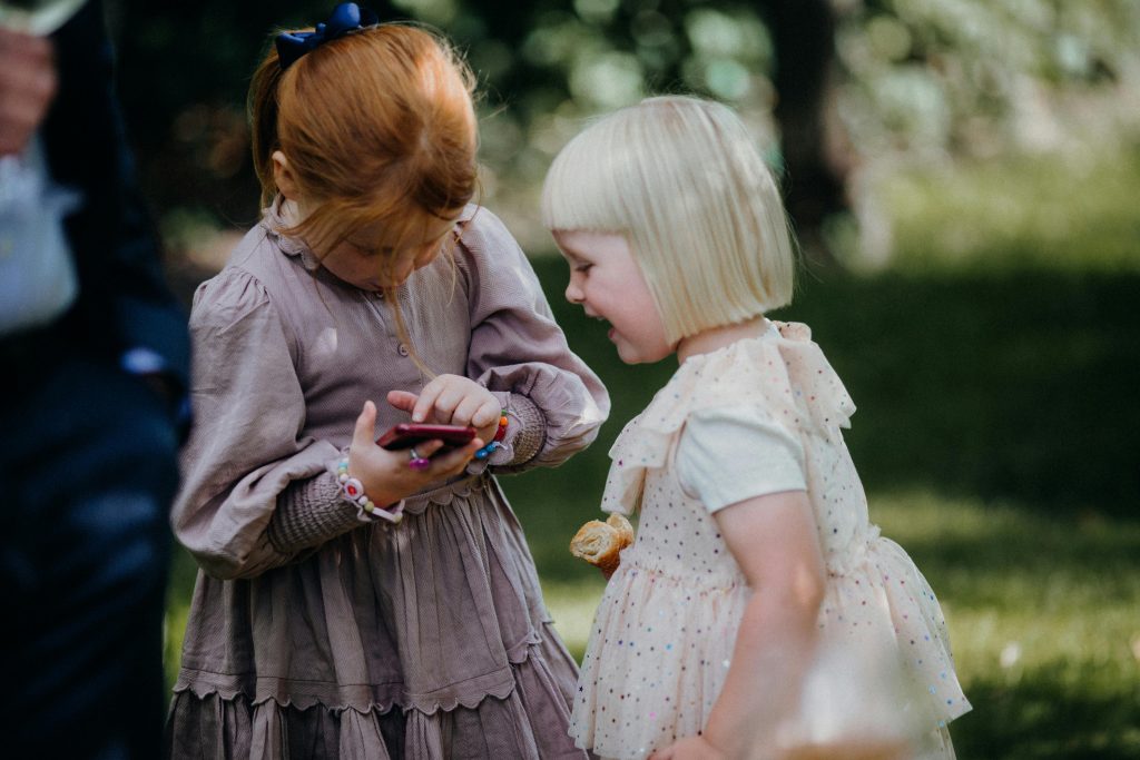 Two young girls with different hair colors are engaged with a smartphone in a sunny outdoor setting.