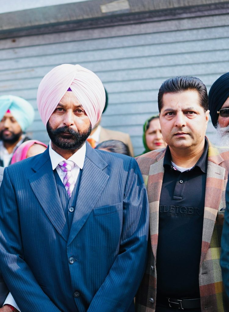 A detailed portrait of two men standing together, one in a turban and suit, the other in modern casual attire.