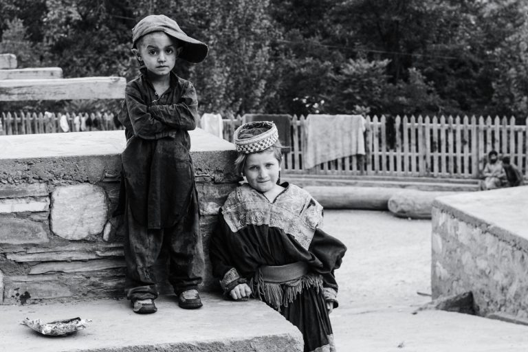 Two children in traditional Chitrali clothing pose outdoors, against a rustic backdrop.