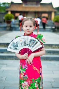 A young girl in a colorful traditional dress holds a decorative fan outdoors.