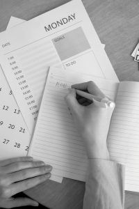 A monochrome image of a person planning their week with a notebook, calendar, and goals list.